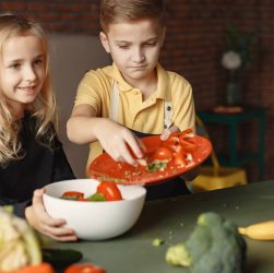 Positive little girl and boy mixing cutted ingredients for salad in big bowl while cooking at table with assorted fresh vegetables and fruits