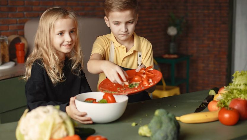 Positive little girl and boy mixing cutted ingredients for salad in big bowl while cooking at table with assorted fresh vegetables and fruits
