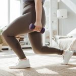 A woman in active wear performs a lunges exercise with dumbbells indoors.