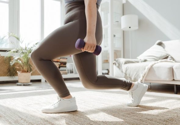 A woman in active wear performs a lunges exercise with dumbbells indoors.
