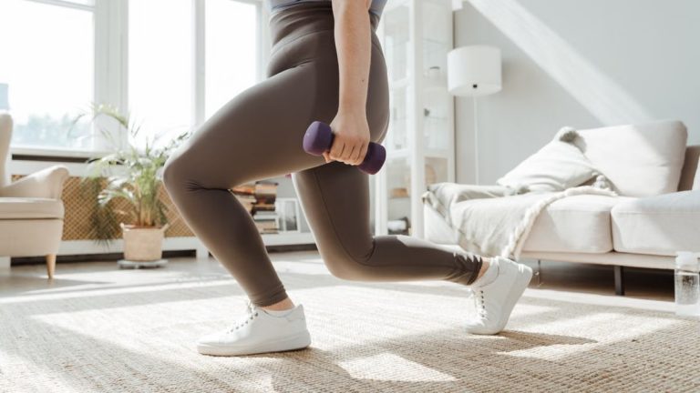 A woman in active wear performs a lunges exercise with dumbbells indoors.